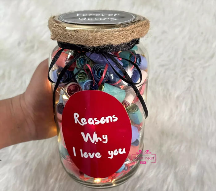 A glass jar filled with colorful rolled paper notes, featuring a red oval label that reads "Reasons Why I Love You," decorated with twine and a black ribbon.